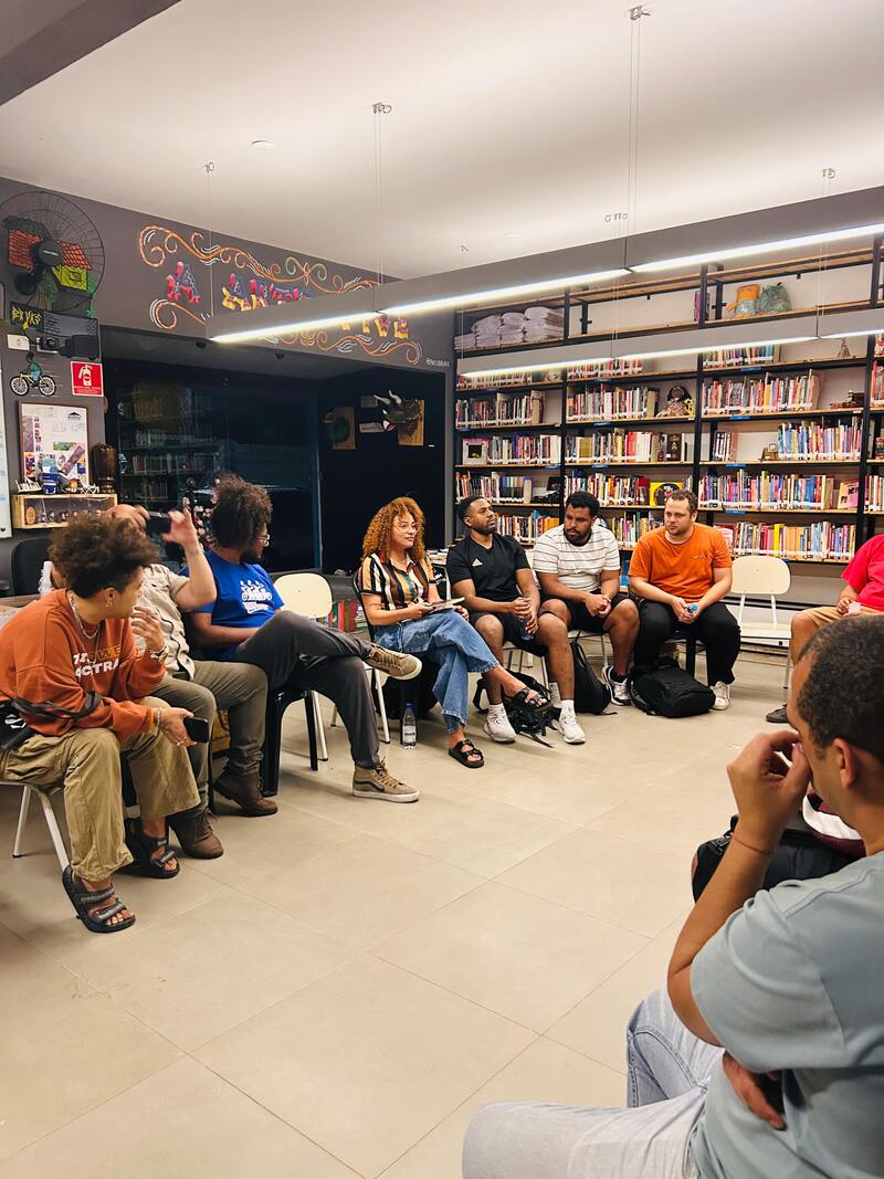 Trata-se de reunião no Ponto de Leitura do Jardim Lapena. É possível ver pessoas sentadas em cadeiras distribuídas pelo espaço. Aparecem ao fundo uma parede na cor preta, assim como uma janela que vai do teto a até quase dois metros de altura, e uma estante que acomoda diversos livros.