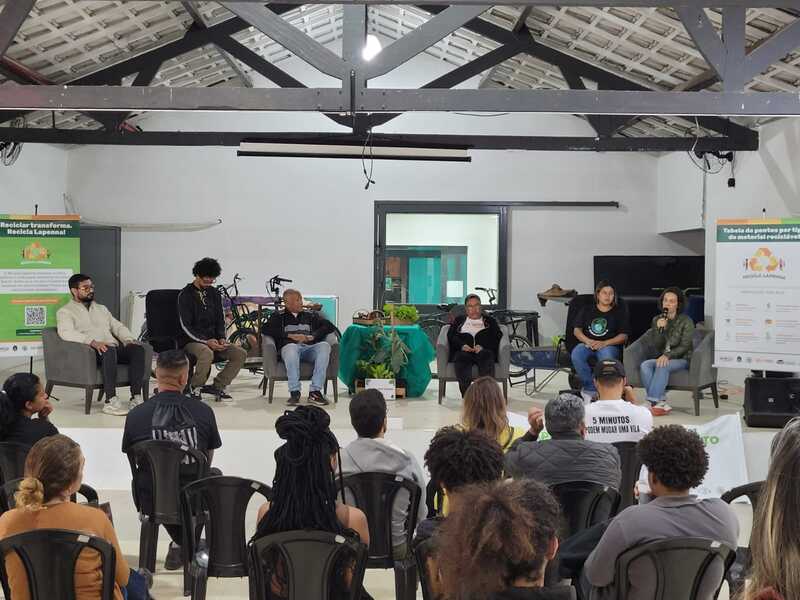 Imagem do evento de lançamento do Recicla Lapenna, no Galpão ZL. Há seis pessoas sentadas no palco do espaço. Há um banner do projeto no canto direito e outro mais para o fundo, à esquerda. Pode-se ver também itens como bicicletas e, ao centro, uma mesa decorada com pano verde e sobre a qual há flores. Há, na parte inferior da foto, pessoas sentadas em cadeiras.