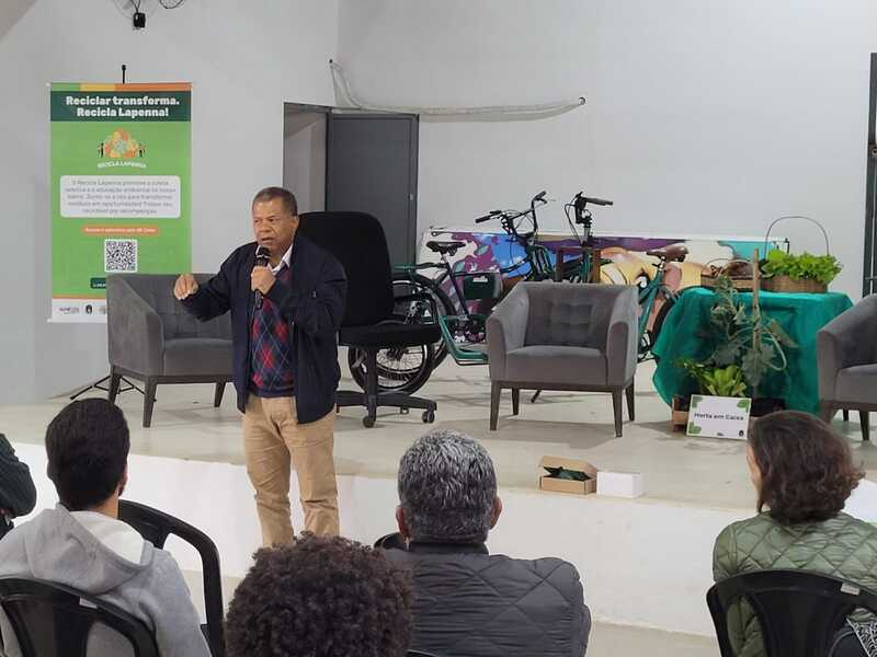 Imagem do evento de lançamento do Recicla Lapenna, no Galpão ZL. Pode-se ver um homem falando ao microfone - ele usa uma blusa com padrão xadrez losangular, uma jaqueta na cor azul e calças da cor bege. Há um banner do projeto ao fundo, à esquerda. Pode-se ver também itens como bicicletas e, ao centro, uma mesa decorada com pano verde e sobre a qual há flores. Há, na parte inferior da foto, pessoas sentadas em cadeiras.