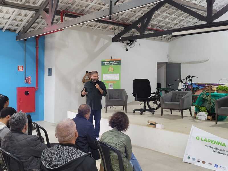 Imagem do evento de lançamento do Recicla Lapenna, no Galpão ZL. Pode-se ver um homem falando ao microfone - trata-se de Marcelo Ribeiro, gerente de Projetos Estratégicos, e ele usa calça jeans e camisa na cor preta. Há um banner do projeto ao fundo, à esquerda. Pode-se ver também itens como bicicletas e, ao centro, uma mesa decorada com pano verde e sobre a qual há flores. Há, na parte inferior da foto, pessoas sentadas em cadeiras.