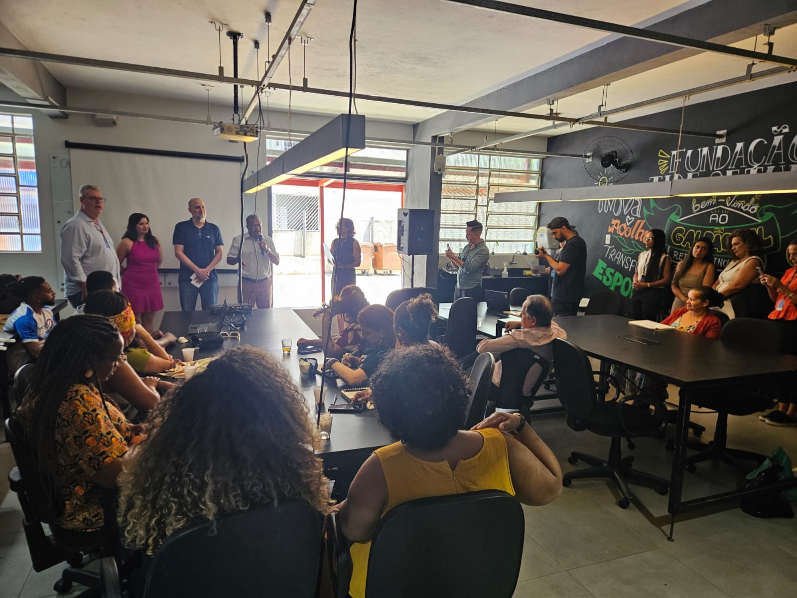 Imagem de reunião de pessoas no espaço coworking do Galpão ZL. Parte do grupo aparece sentado em frente a uma das mesas do equipamento, enquanto outras estão em pé em pontos diversos do espaço - e da foto. Ao centro, um homem aparece falando ao microfone durante atividade do projeto Sebrae Aqui.