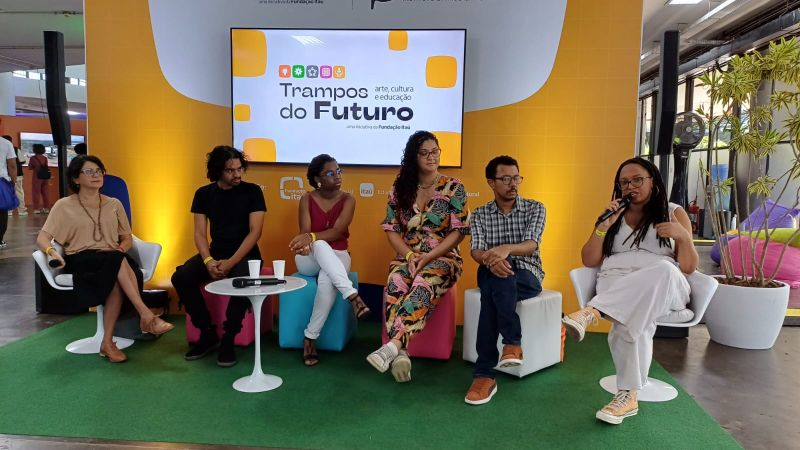 Participantes do evento Trampos do Futuro sentados em pufes coloridos, com Natália Carneiro falando ao microfone.