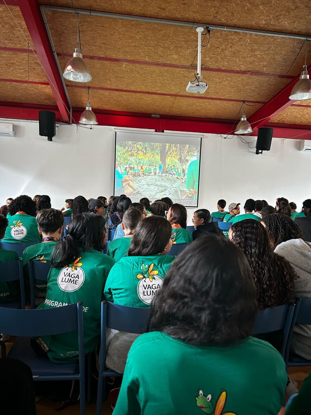 Imagem de reunião da Rede Vaga Lume. As crianças e adolescentes participantes aparecem de costas, com camisetas do projeto, sentadas em cadeiras dispostas em um salão.