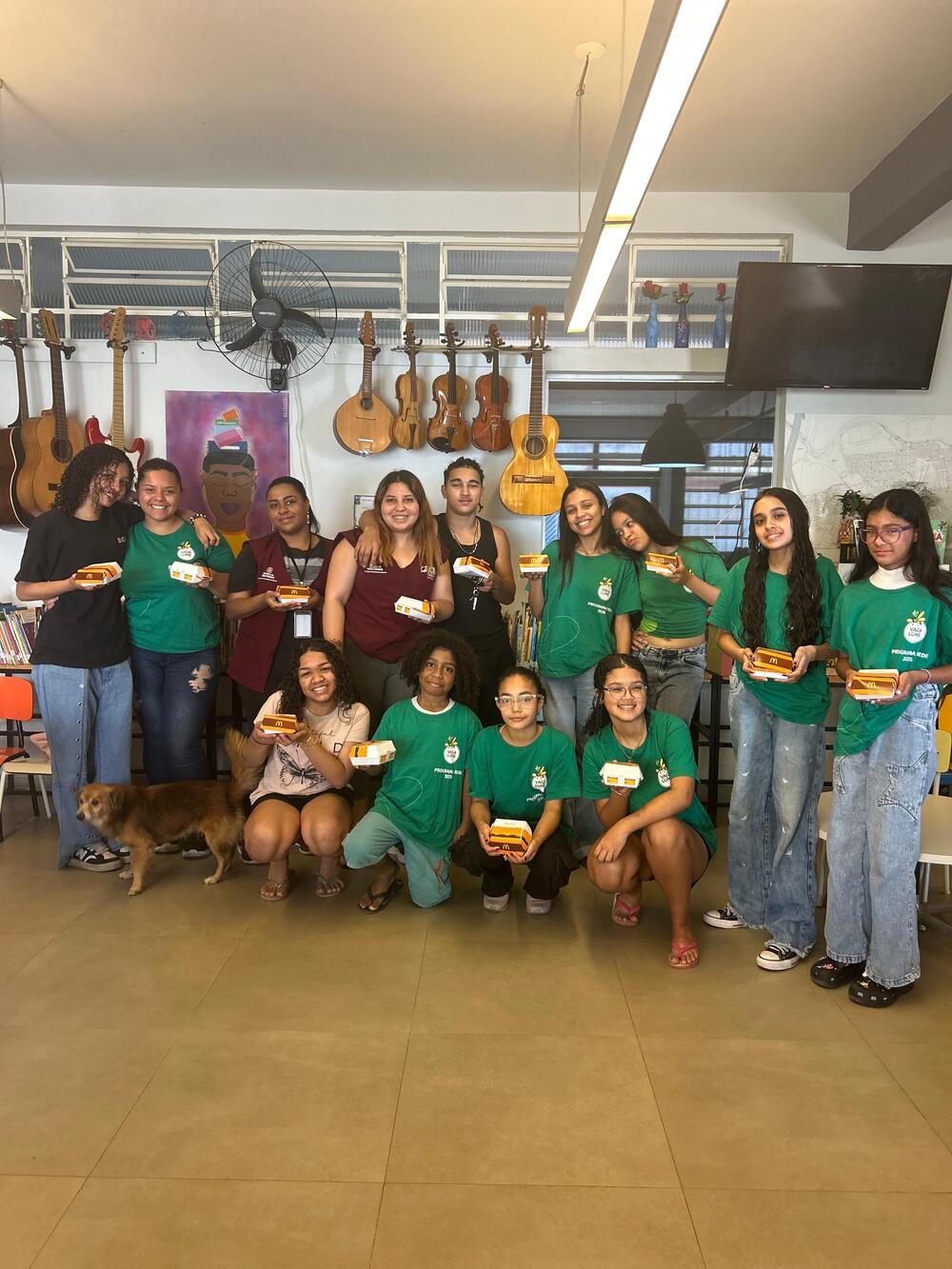 Imagem do Ponto de Leitura do Jardim Lapenna. É possível ver crianças e adolescentes integrantes da Rede Vaga Lume - elas usam camisetas verdes do projeto. Nove entre elas aparecem em pé, enquanto outras quatro pessoas estão agachadas. É possível ver instrumentos de corda - violões e violinos - e um quadro roxo na parede ao fundo.