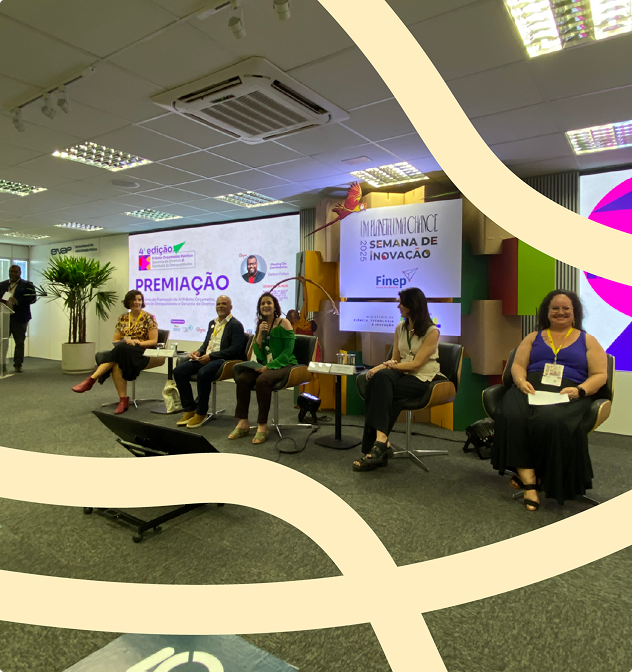 Imagem do evento de premiação do IV Prêmio Orçamento Público, Garantia de Direitos e Combate às Desigualdades na qual aparecem cinco pessoas sentadas e uma no canto esquerdo, em pé e em frente a um púlpito transparente.